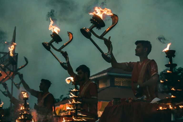 Evening Ganga Aarti ceremony on the banks of the Ganges River in Varanasi, with priests holding lit oil lamps and a vibrant crowd in attendance.