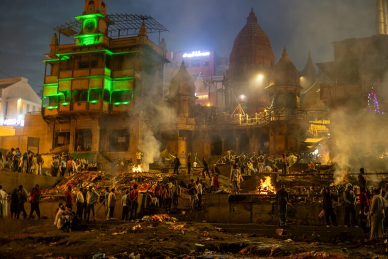 Manikarnika Ghat in Varanasi, India, with smoke rising from funeral pyres along the Ganges River, surrounded by ancient buildings and a somber atmosphere.