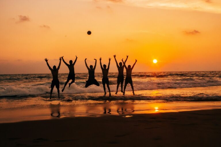 Group of friends jumping in excitement against a stunning beach sunset, with the orange sky and sun reflecting on the water and wet sand