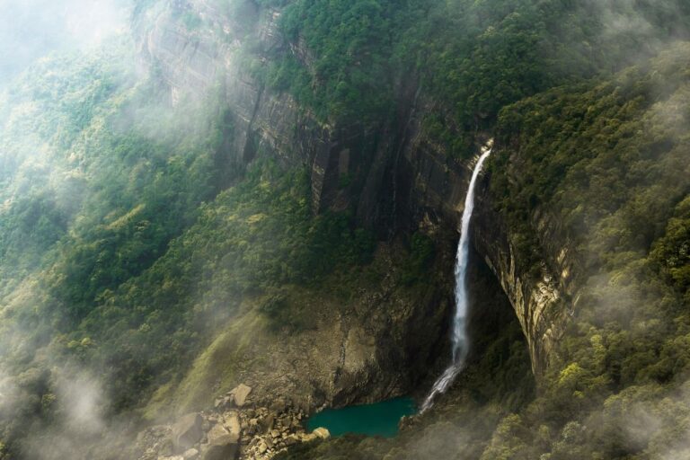 Majestic waterfall cascading into a turquoise pool, surrounded by misty, lush green cliffs in Meghalaya, India.